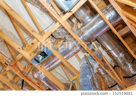 Workers install ventilation ducts between wooden beams in home under construction, utilizing tools ladders. 132559081