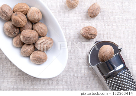 Whole nutmegs, in white bowl and with a nutmeg grater on the right side, on linen. Dried seeds of fragrant or true nutmeg fruits, Myristica fragrans. Used as spice, and for their psychoactive effects. 132563414