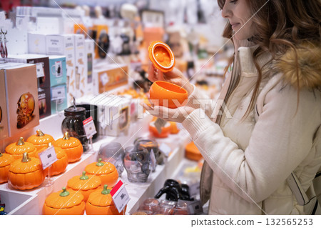 On the eve of Halloween, a woman browses a local market, selecting colorful ceramic pumpkins and unique jars for home decor. She enjoys the festive shopping atmosphere. On the eve of Halloween, a woman browses a local market, selecting colorful ceramic pumpkins and unique jars for home decor. She enjoys the festive shopping atmosphere. 132565253