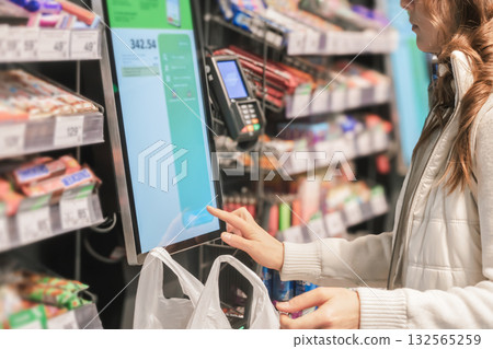 A woman pays for purchases and groceries in a modern store at a self-service checkout with a touchscreen, close-up. A woman pays for purchases and groceries in a modern store at a self-service checkout with a touchscreen, close-up. 132565259