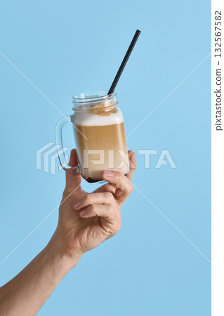 Hand holding glass mug with iced latte and straw on blue background for cafe ads 132567582
