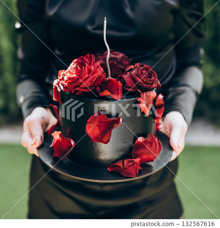 Unique black cake decorated with real rose buds and petals, held by a woman without face visible, symbolizing stylish and elegant celebration. 132567616