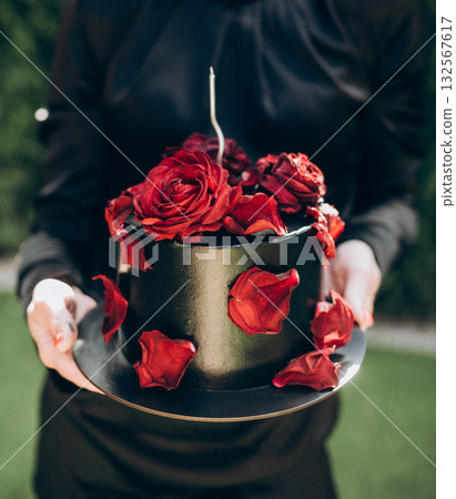Unique black cake decorated with real rose buds and petals, held by a woman without face visible, symbolizing stylish and elegant celebration. 132567617