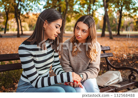 Two girls sit closely on a park bench surrounded by vibrant autumn leaves Two girls sit closely on a park bench surrounded by vibrant autumn leaves 132568488