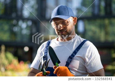Professional gardener in uniform holding tools while standing in green garden Professional gardener in uniform holding tools while standing in green garden 132568541