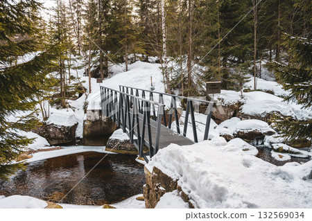 A picturesque winter scene showcases a creek in the Czech Republic known as Jizerka. Snow blankets the ground, and a bridge spans the cold waters, creating a tranquil atmosphere. A picturesque winter scene showcases a creek in the Czech Republic known as Jizerka. Snow blankets the ground, and a bridge spans the cold waters, creating a tranquil atmosphere. 132569034