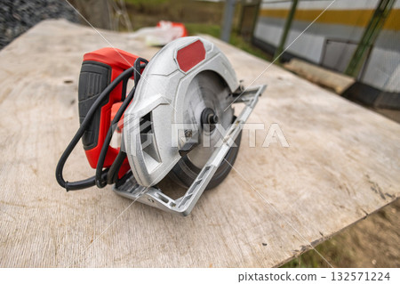 A circular saw sits idle on a wooden surface at a construction site, showcasing its metallic blade and red handle against a blurred background of equipment A circular saw sits idle on a wooden surface at a construction site, showcasing its metallic blade and red handle against a blurred background of equipment 132571224