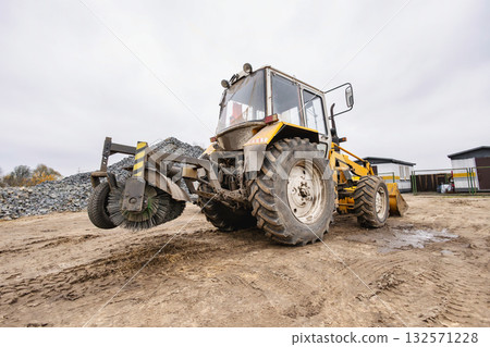 A large tractor equipped with a sweeping attachment is parked at a construction site, surrounded by piles of gravel on a cloudy day in autumn 132571228