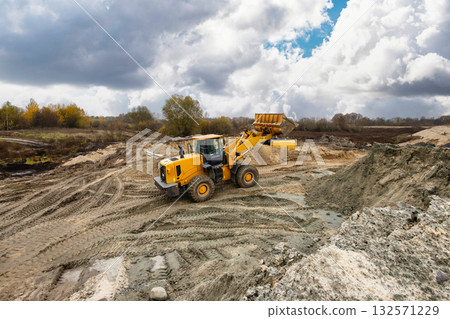 A yellow loader operates on a construction site, moving dirt and gravel while clouds gather overhead in a remote area during daytime A yellow loader operates on a construction site, moving dirt and gravel while clouds gather overhead in a remote area during daytime 132571229