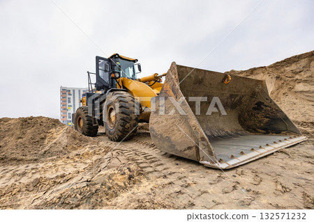 A large loader works at a construction site, moving soil with a bucket in front of a building in an urban area on a cloudy day. A large loader works at a construction site, moving soil with a bucket in front of a building in an urban area on a cloudy day. 132571232