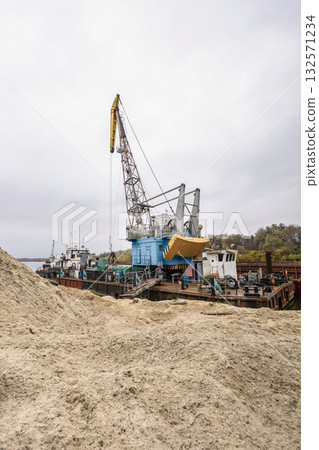 A large crane is positioned beside a river, carefully lifting materials at a construction site. The day is overcast, reflecting autumn's chill A large crane is positioned beside a river, carefully lifting materials at a construction site. The day is overcast, reflecting autumn's chill 132571234