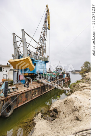A large crane is positioned on a barge at a river construction site, with equipment lined up for operation on a cloudy day A large crane is positioned on a barge at a river construction site, with equipment lined up for operation on a cloudy day 132571237
