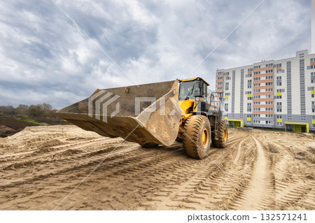 A large yellow loader is lifting dirt at a construction site near a new residential building under a cloudy sky, demonstrating active site work A large yellow loader is lifting dirt at a construction site near a new residential building under a cloudy sky, demonstrating active site work 132571241