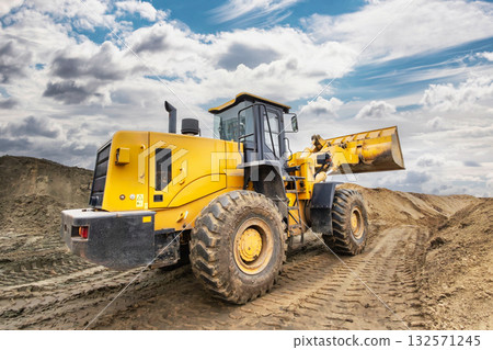 A yellow loader works on a construction site, moving earth and building materials, with clouds in the sky and dirt surrounding the area 132571245