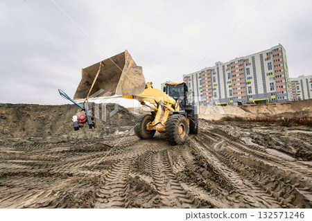 A large yellow loader works at a construction site, moving dirt near apartment buildings on a cloudy day. The ground is uneven with tire tracks visible A large yellow loader works at a construction site, moving dirt near apartment buildings on a cloudy day. The ground is uneven with tire tracks visible 132571246