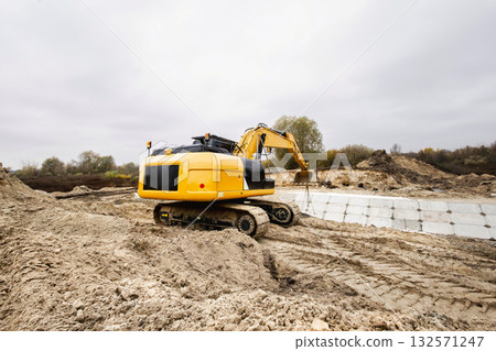 Construction site features an excavator digging in soft earth under a gray sky. Surrounding area shows autumn trees, creating a tranquil scene 132571247