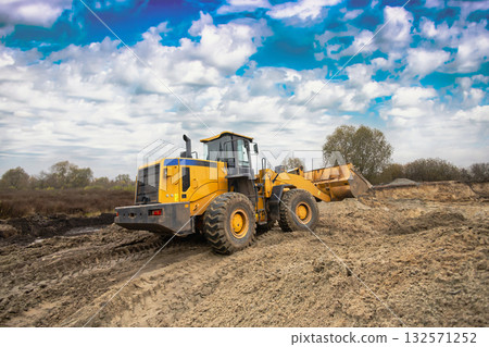 A large yellow loader moves dirt across a construction site under a cloudy sky. Trees are visible in the background, adding to the natural setting A large yellow loader moves dirt across a construction site under a cloudy sky. Trees are visible in the background, adding to the natural setting 132571252