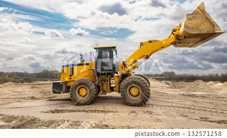 A yellow loader works on a construction site, lifting sand and gravel while surrounded by piles of material under a partly cloudy sky 132571258