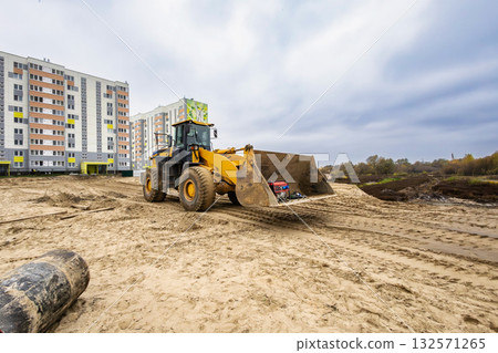 A large construction vehicle works on sandy terrain close to apartment buildings while the sky remains cloudy, indicating a cool day A large construction vehicle works on sandy terrain close to apartment buildings while the sky remains cloudy, indicating a cool day 132571265