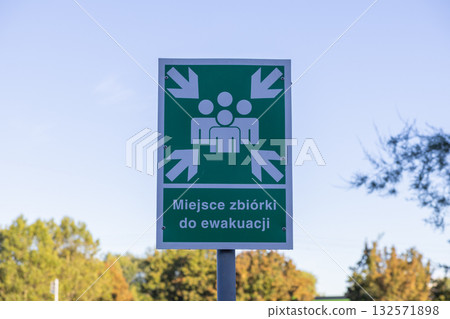 Green emergency evacuation assembly point sign, clearly visible outdoors, featuring people and arrows symbol, indicating a public safety and crisis management location 132571898