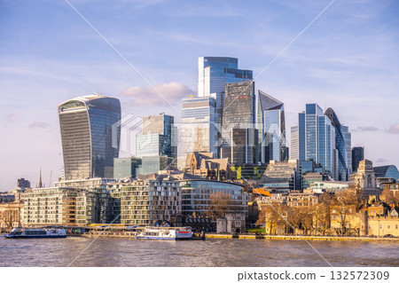 Modern skyscrapers rise against a clear sky in London City, reflecting on the River Thames. Boats glide along the water, showcasing the vibrant urban landscape of England. 132572309