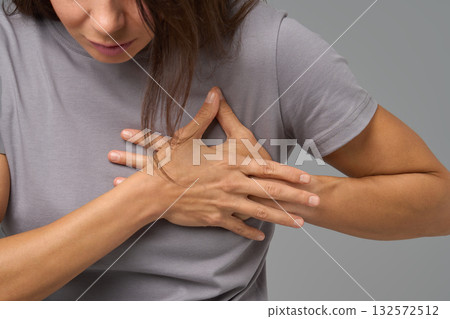 Woman clutching chest in discomfort, close-up studio view on neutral grey background, Woman clutching chest in discomfort, close-up studio view on neutral grey background, 132572512
