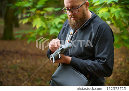 A focused man adjusts his drone while surrounded by vibrant trees displaying shades of green and gold. It is a peaceful afternoon in nature. A focused man adjusts his drone while surrounded by vibrant trees displaying shades of green and gold. It is a peaceful afternoon in nature. 132572725