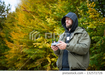 A man in a hoodie controls a drone with a remote as golden and orange leaves surround him in a serene park. The atmosphere is calm and filled with nature's beauty. A man in a hoodie controls a drone with a remote as golden and orange leaves surround him in a serene park. The atmosphere is calm and filled with nature's beauty. 132572726