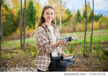 In a beautiful forest during autumn, a smiling young woman stands proudly holding a drone. Colorful trees surround her, showcasing the season's vibrant hues. 132572732