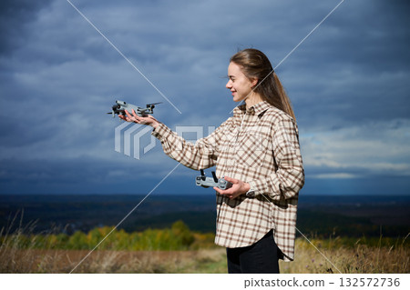 A young woman stands in a grassy area, holding drones in both hands as she prepares to fly them. The clouds create a moody backdrop, enhancing the adventurous scene. 132572736