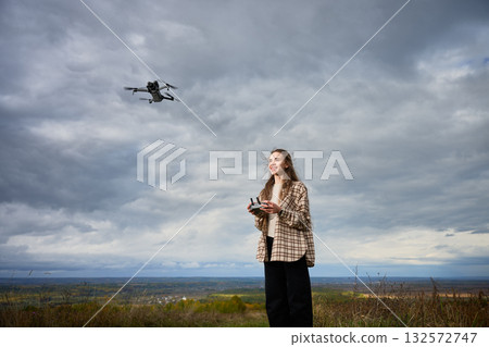 A young woman stands on a hilltop with a remote control, flying her drone above an expansive vista. The clouds create a dramatic backdrop as she smiles joyfully. 132572747