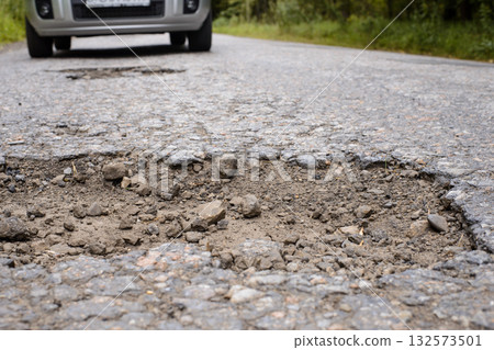 Big pothole in asphalt road and modern car driving right to it in the background 132573501