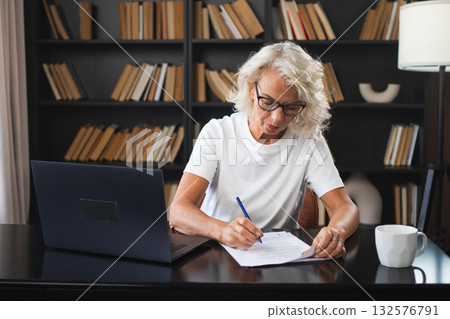 Middle aged senior woman using laptop computer writing notes at workplace. Focused mature old businesswoman work at office making written records doing online work. Mature boss professional worker 132576791