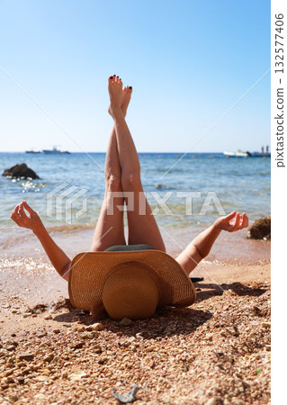 A woman in a wide sun hat lies on the sand overlooking the sea with her legs raised to the sky, enjoying the sun's rays A woman in a wide sun hat lies on the sand overlooking the sea with her legs raised to the sky, enjoying the sun's rays 132577406