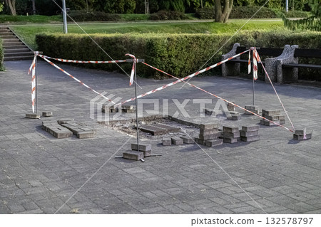 Public park paved walkway under renovation, marked with red and white warning tape around an exposed drain and stacked paving stones 132578797