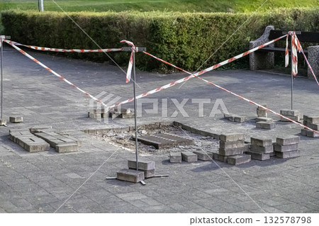 Paved urban walkway under repair with removed bricks surrounding a storm drain, safely cordoned off by red and white warning tape, indicating maintenance work Paved urban walkway under repair with removed bricks surrounding a storm drain, safely cordoned off by red and white warning tape, indicating maintenance work 132578798