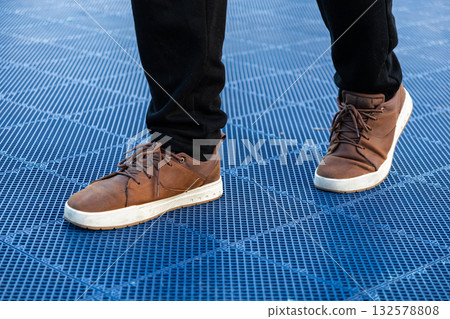 Person's legs in black pants and brown leather sneakers with white soles standing on a vibrant blue grid textured outdoor surface, emphasizing urban casual footwear Person's legs in black pants and brown leather sneakers with white soles standing on a vibrant blue grid textured outdoor surface, emphasizing urban casual footwear 132578808