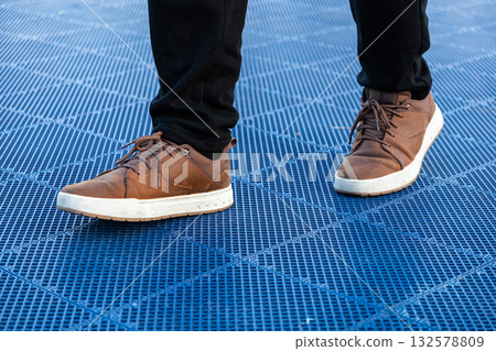 Person's feet wearing comfortable brown leather casual shoes with white soles, standing on a vibrant blue grid surface, highlighting modern urban outdoor fashion Person's feet wearing comfortable brown leather casual shoes with white soles, standing on a vibrant blue grid surface, highlighting modern urban outdoor fashion 132578809