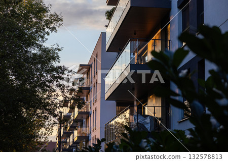 Modern apartment building facade showcases sleek glass balconies reflecting warm golden hour sunlight, highlighting contemporary urban living and residential architecture design 132578813