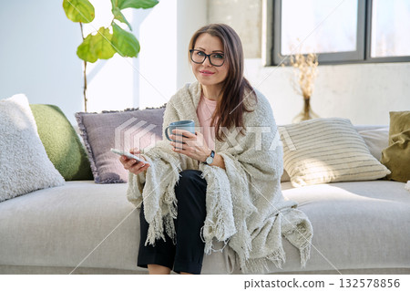Woman under blanket with mug of hot drink using mobile phone sitting on sofa 132578856