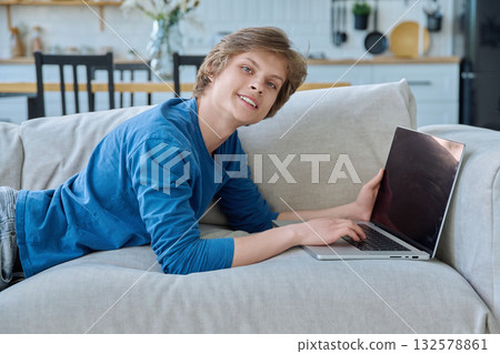Handsome teenage boy lying at home on couch with laptop, looking at camera Handsome teenage boy lying at home on couch with laptop, looking at camera 132578861