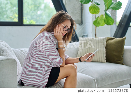 Sad serious middle aged woman with smartphone sitting on sofa 132578878