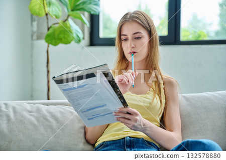 Young student girl study online, sitting at home on couch with textbook Young student girl study online, sitting at home on couch with textbook 132578880