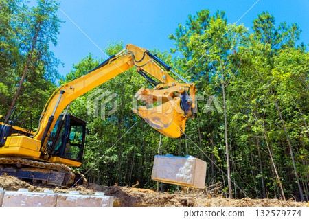 Excavator operates in laying large stone block during construction retaining wall landscaping work 132579774