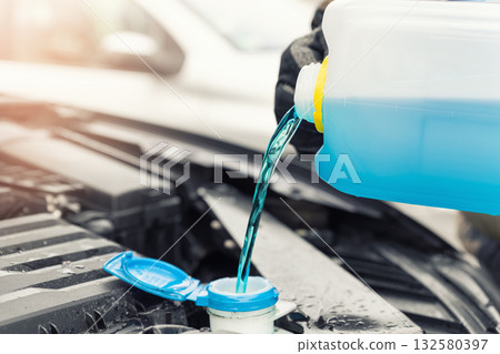 Close-up detail of driver or mechanic hand in gloves pouring blue antifreeze liquid for winter car windshield screen washing with bootle watering can. Car seasonal service and maintenance concept 132580397