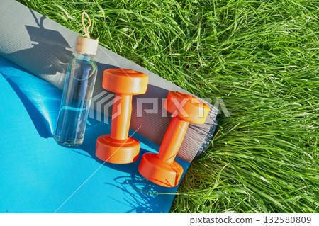 Outdoor fitness setup with orange dumbbells, yoga mat, and water bottle on grass 132580809