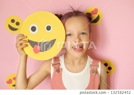 A funny, humorous girl sticks out tongue, holding a grimacing smiley face in hands. World Emoji and Social Media Day. 132582452