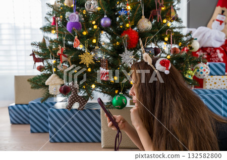 Young woman with long hair using smartphone near decorated Christmas tree with glowing lights Young woman with long hair using smartphone near decorated Christmas tree with glowing lights 132582800