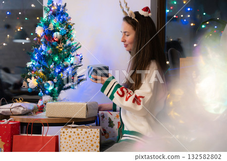 Young woman with reindeer headband holding gift box beside decorated tree at home Young woman with reindeer headband holding gift box beside decorated tree at home 132582802