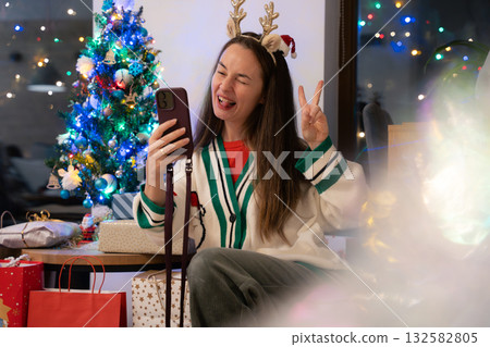 Playful young woman taking funny Christmas selfie at home near decorated tree with festive lights Playful young woman taking funny Christmas selfie at home near decorated tree with festive lights 132582805